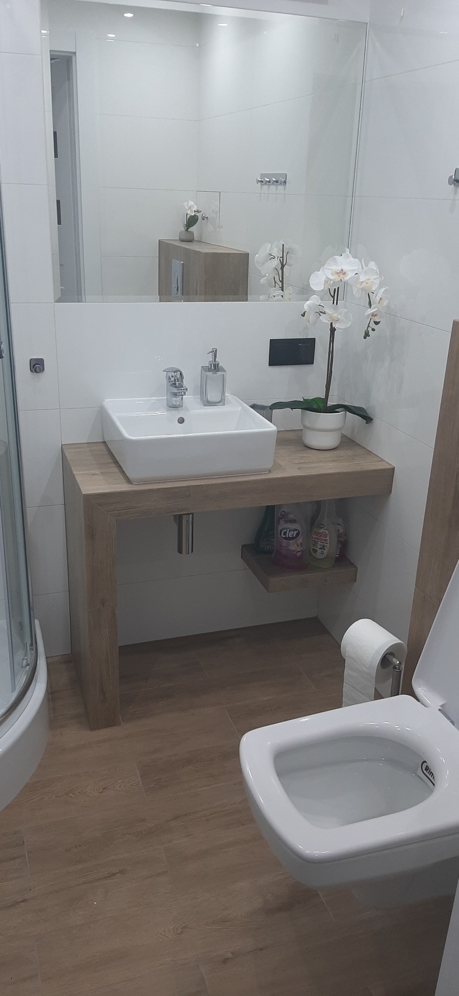 Nowoczesna łazienka z wiszącą toaletą, umywalką nablatową na drewnopodobnym blacie, dużym lustrem i białymi płytkami, uzupełniona dekoracyjną orchideą w doniczce.