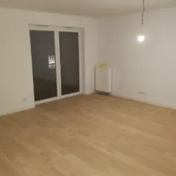 Jasne panele podłogowe w pustym pokoju z białymi ścianami, grzejnikiem z żółtą ścierką i wiszącą żarówką, widok na przesuwne drzwi balkonowe.