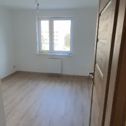 Pusty pokój z panelami podłogowymi w odcieniach szarości, białymi ścianami i kaloryferem pod oknem, widok zza otwartych drewnianych drzwi z poziomymi intarsjami.