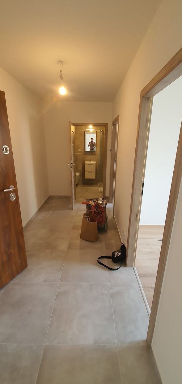 Wyremontowany korytarz z szarymi płytkami podłogowymi i drewnianymi ościeżnicami, widok na łazienkę z lustrem, w tle narzędzia i torba robocza.