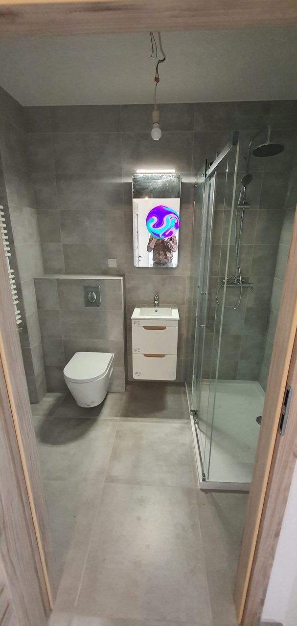 Nowoczesna łazienka z szarymi płytkami, kabiną prysznicową, podwieszaną toaletą i szafką z umywalką, widoczne niedokończone podłączenie oświetlenia sufitowego.