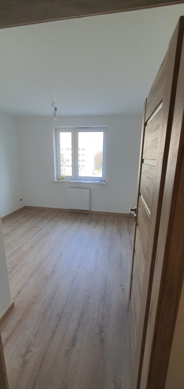 Pusty pokój z panelami podłogowymi w odcieniach szarości, białymi ścianami i kaloryferem pod oknem, widok zza otwartych drewnianych drzwi z poziomymi intarsjami.