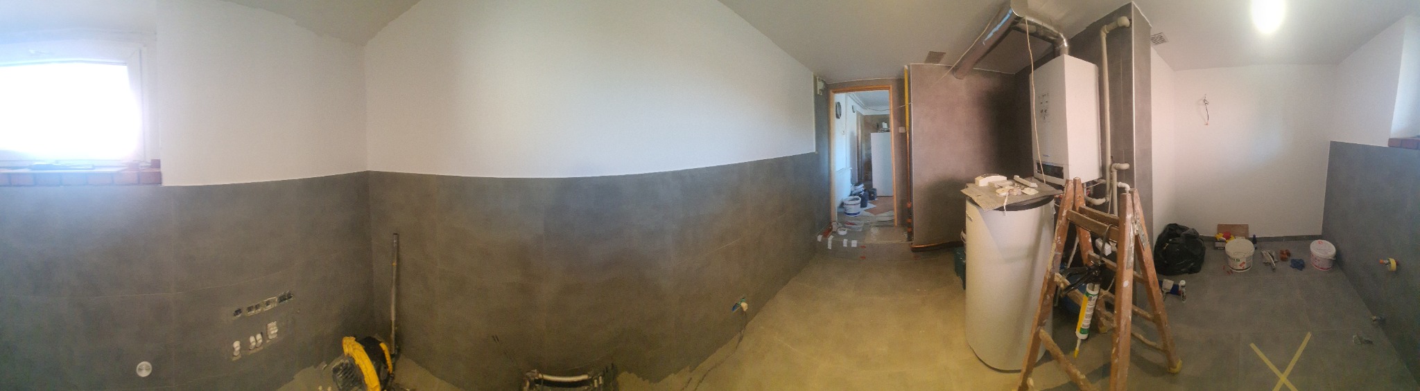 Panoramiczny widok wnętrza w trakcie prac wykończeniowych: szare płytki do połowy ściany, białe ściany powyżej, widoczny bojler, drabina, narzędzia i materiały budowlane, przygotowane do dalszych...