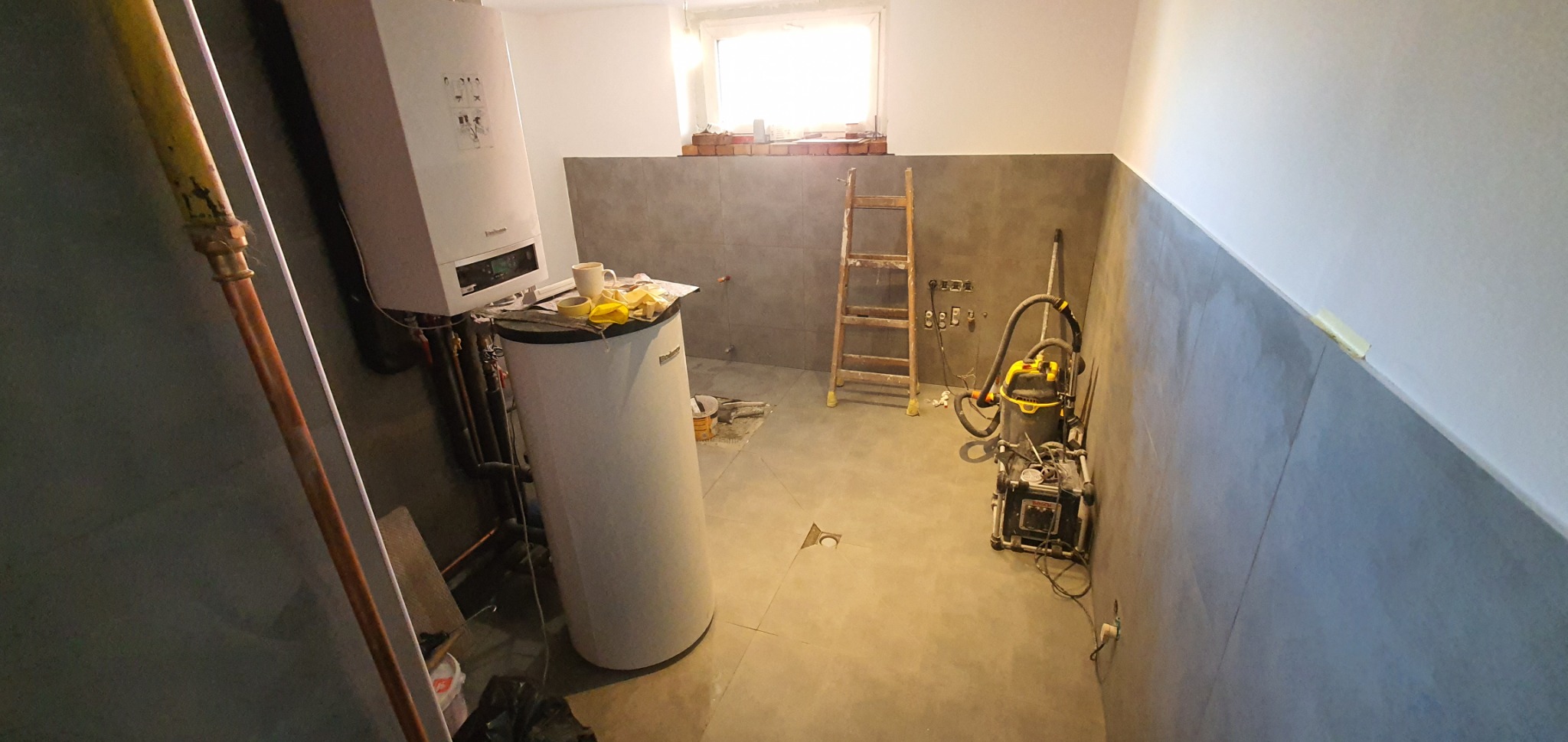 Łazienka w trakcie prac wykończeniowych: częściowo położone szare płytki, widoczny bojler, piec grzewczy, drabina, odkurzacz przemysłowy i narzędzia.
