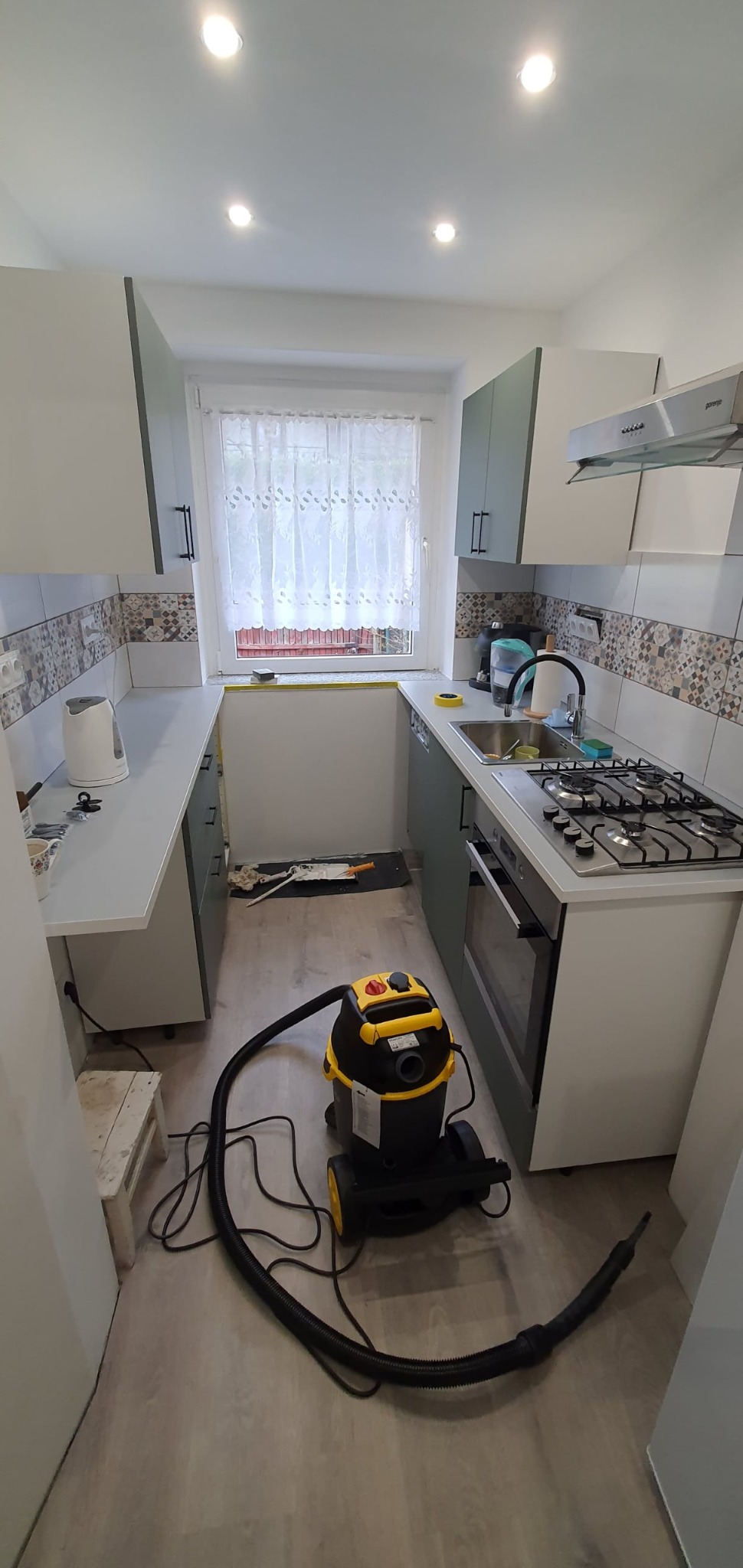 Wąska kuchnia z jasnymi szafkami, płytkami mozaikowymi nad blatem i oknem z firanką. Na podłodze stoi odkurzacz przemysłowy.