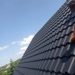 Nowy, ciemny dach z blachodachówki, widoczny z boku pod kątem, z fragmentem postaci dekarza w prawym górnym rogu i błękitnym niebem w tle.