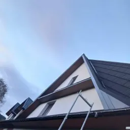 Dwuspadowy dach z ciemnej blachy trapezowej na budynku z ceglaną podstawą, widoczna drabina oparta o ścianę.