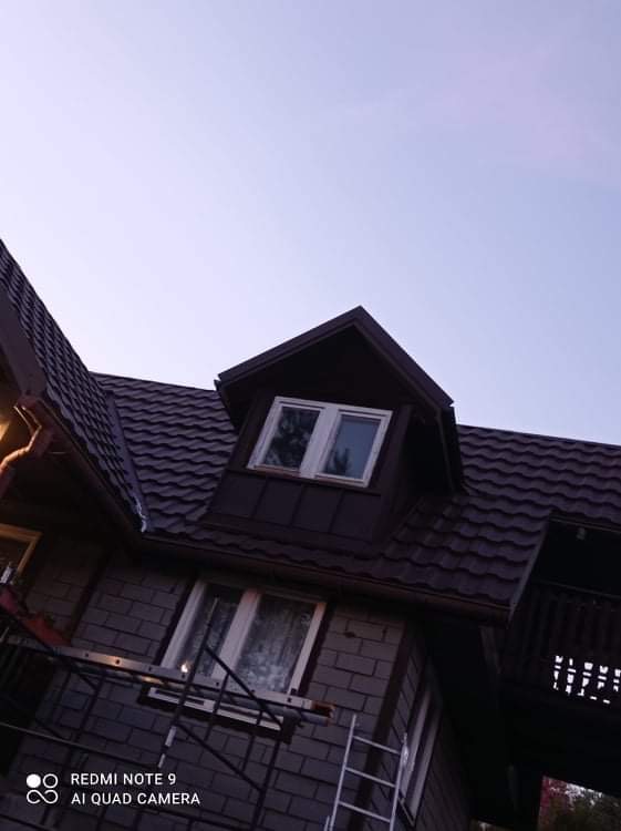 Dom z ciemnobrązowym dachem z blachodachówki, widoczne okno dachowe i drabina oparta o ścianę, niebieskie niebo w tle.