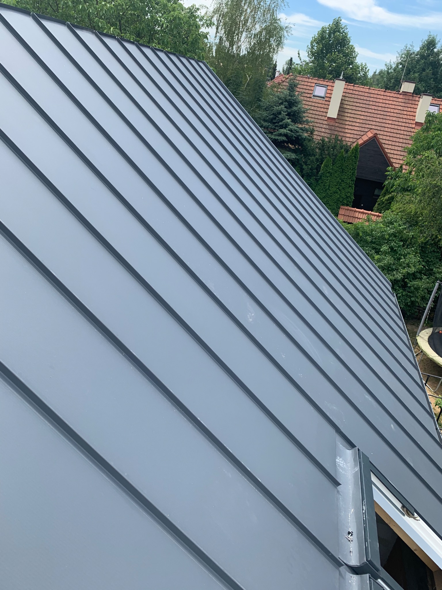 Nowy, szary dach z blachy na domu jednorodzinnym, widok z góry, w tle zieleń drzew i fragment dachu sąsiedniego budynku z czerwonej dachówki.