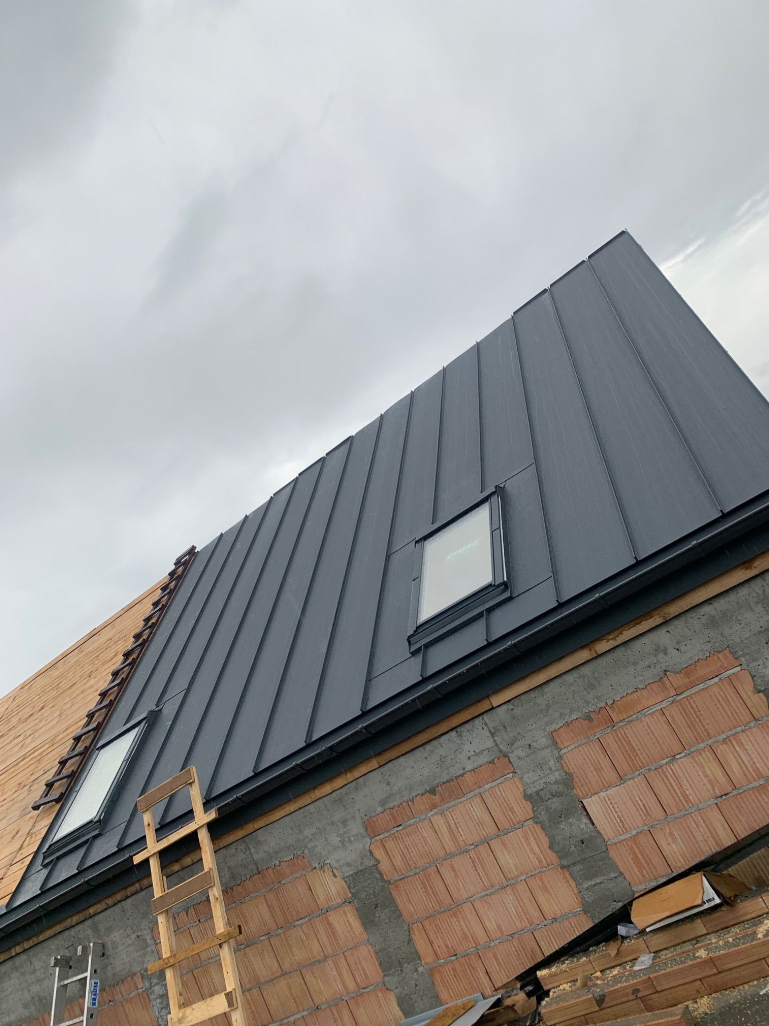 Częściowo pokryty blachą dach z oknami dachowymi, widoczne ceglane ściany i drabiny na tle pochmurnego nieba.