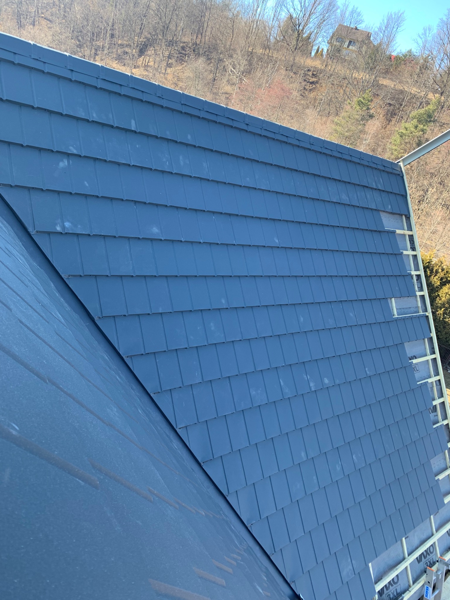 Nowy, grafitowy dach z dachówki bitumicznej w trakcie montażu, widoczne łaty i kontrłaty, w tle zalesione wzgórze z domem.