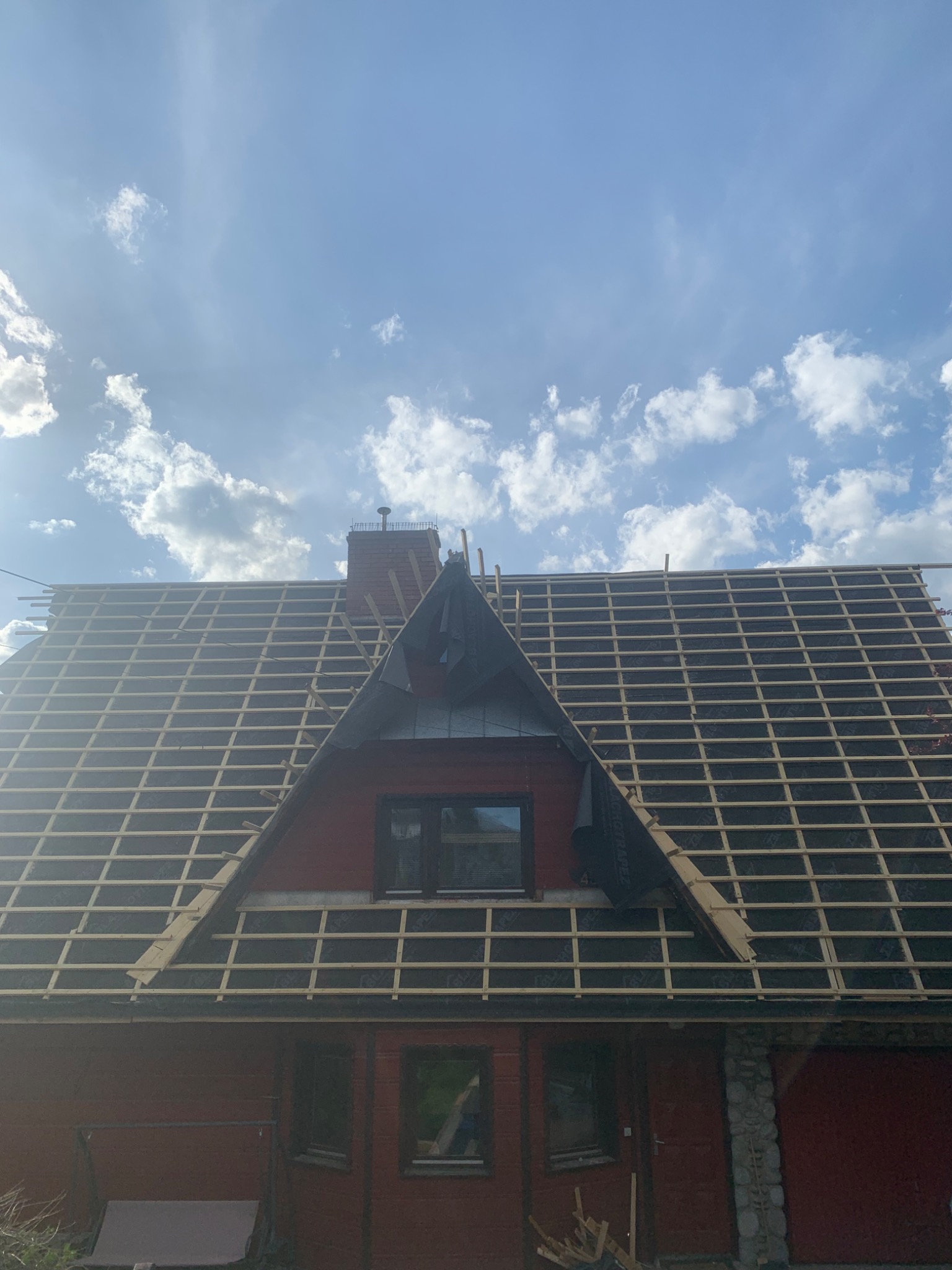 Dom z częściowo zdjętym starym pokryciem dachowym, widoczna więźba i membrana dachowa, przygotowany do położenia nowego dachu, na tle błękitnego nieba z chmurami.