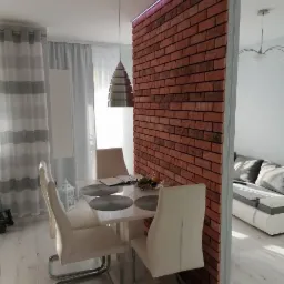 Jasna jadalnia z białym stołem i krzesłami, oddzielona od salonu ścianką z cegieł z podświetleniem LED.