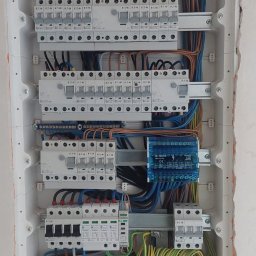 ELEKTRO-M.A.K.Z. - Rozdzielnica elektryczna w białej obudowie, widoczne zabezpieczenia, przewody w różnych kolorach, listwy zaciskowe. Ściana wokół rozdzielnicy wymaga wykończenia.