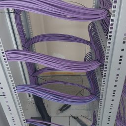 ELEKTRO-M.A.K.Z. - Widok z dołu na uporządkowane fioletowe przewody telekomunikacyjne w metalowej szafie serwerowej, z widocznymi narzędziami i czarną rurą osłonową.