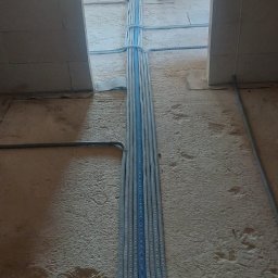 ELEKTRO-M.A.K.Z. - Rury instalacyjne, szare i niebieskie, prowadzone po surowym betonowym podłożu wewnątrz budynku w stanie deweloperskim, zabezpieczone opaskami.