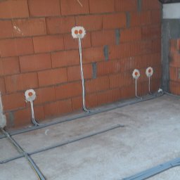 ELEKTRO-M.A.K.Z. - Surowa ściana z czerwonej cegły dziurawki z widocznymi puszkami elektrycznymi i przewodami, szare rury karbowane na podłodze, poddasze w stanie deweloperskim.