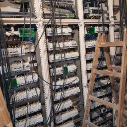 Rozbudowana rozdzielnica elektryczna z licznymi zabezpieczeniami i okablowaniem, widoczna drabina monterska.