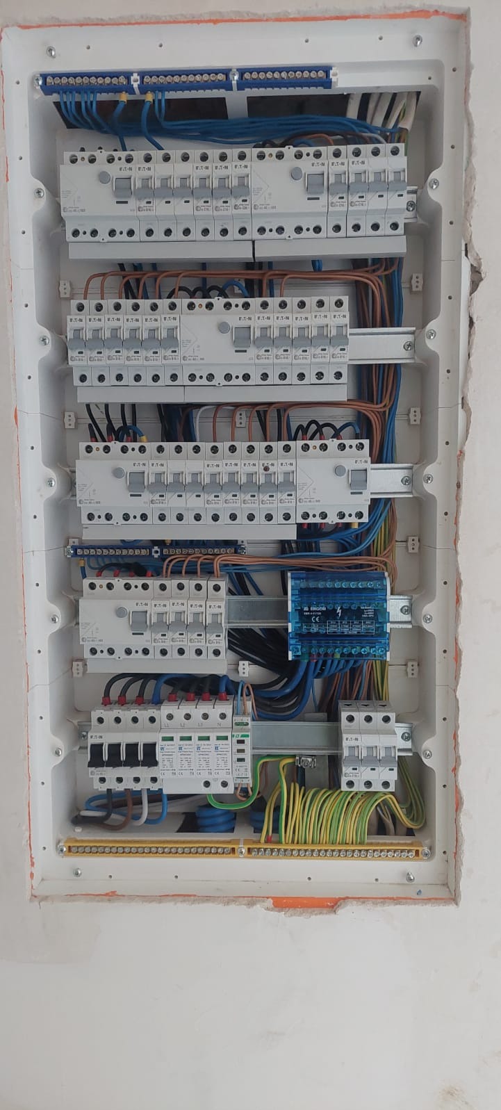 Rozdzielnica elektryczna w białej obudowie, widoczne zabezpieczenia, przewody w różnych kolorach, listwy zaciskowe. Ściana wokół rozdzielnicy wymaga wykończenia.