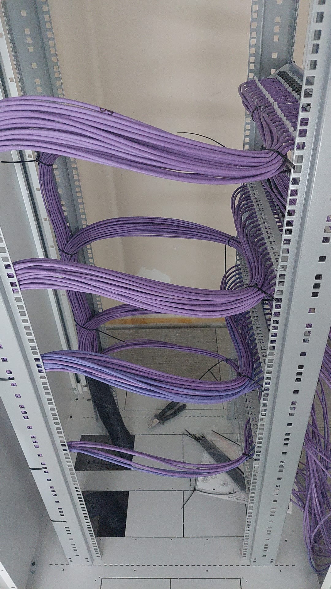 Widok z dołu na uporządkowane fioletowe przewody telekomunikacyjne w metalowej szafie serwerowej, z widocznymi narzędziami i czarną rurą osłonową.