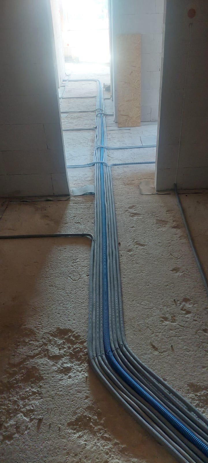 Rury instalacyjne, szare i niebieskie, prowadzone po surowym betonowym podłożu wewnątrz budynku w stanie deweloperskim, zabezpieczone opaskami.