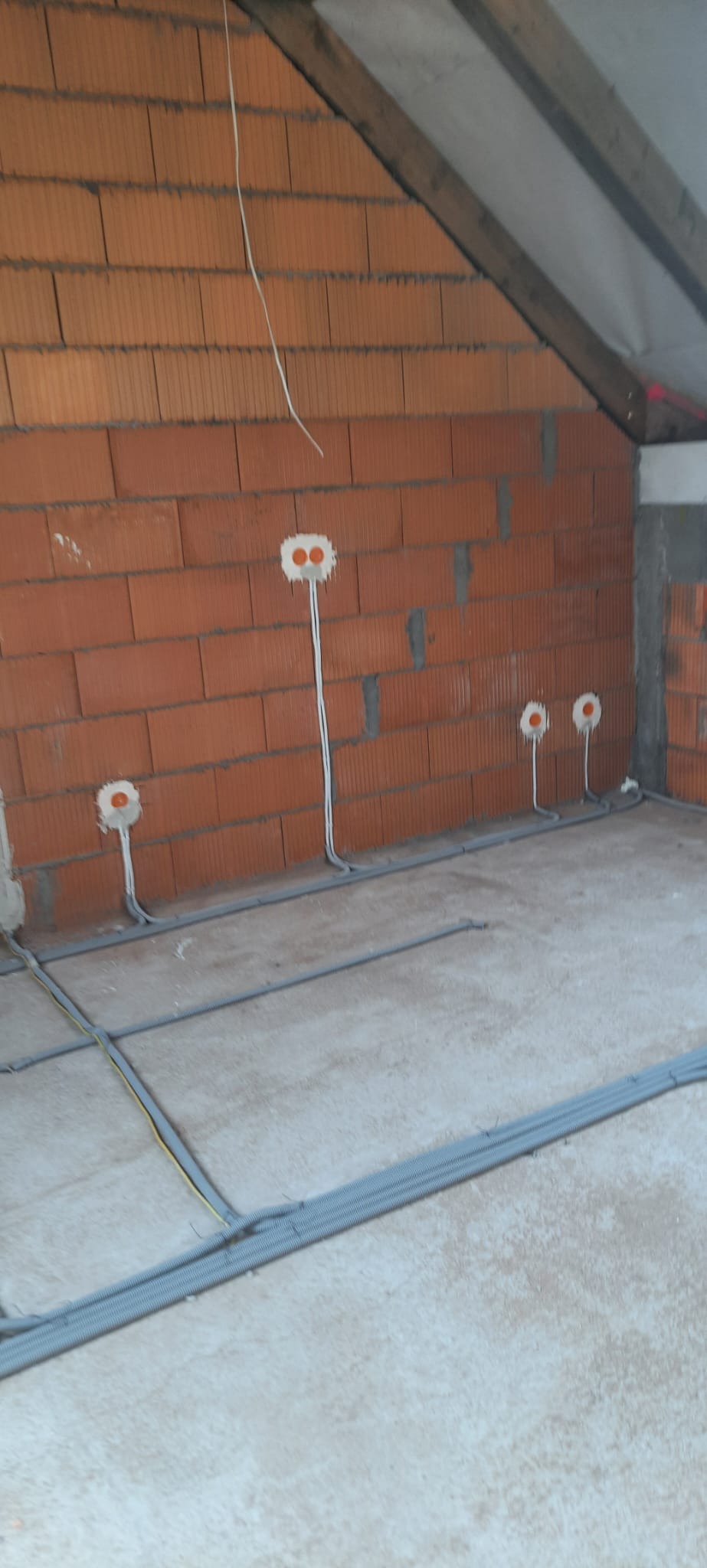 Surowa ściana z czerwonej cegły dziurawki z widocznymi puszkami elektrycznymi i przewodami, szare rury karbowane na podłodze, poddasze w stanie deweloperskim.