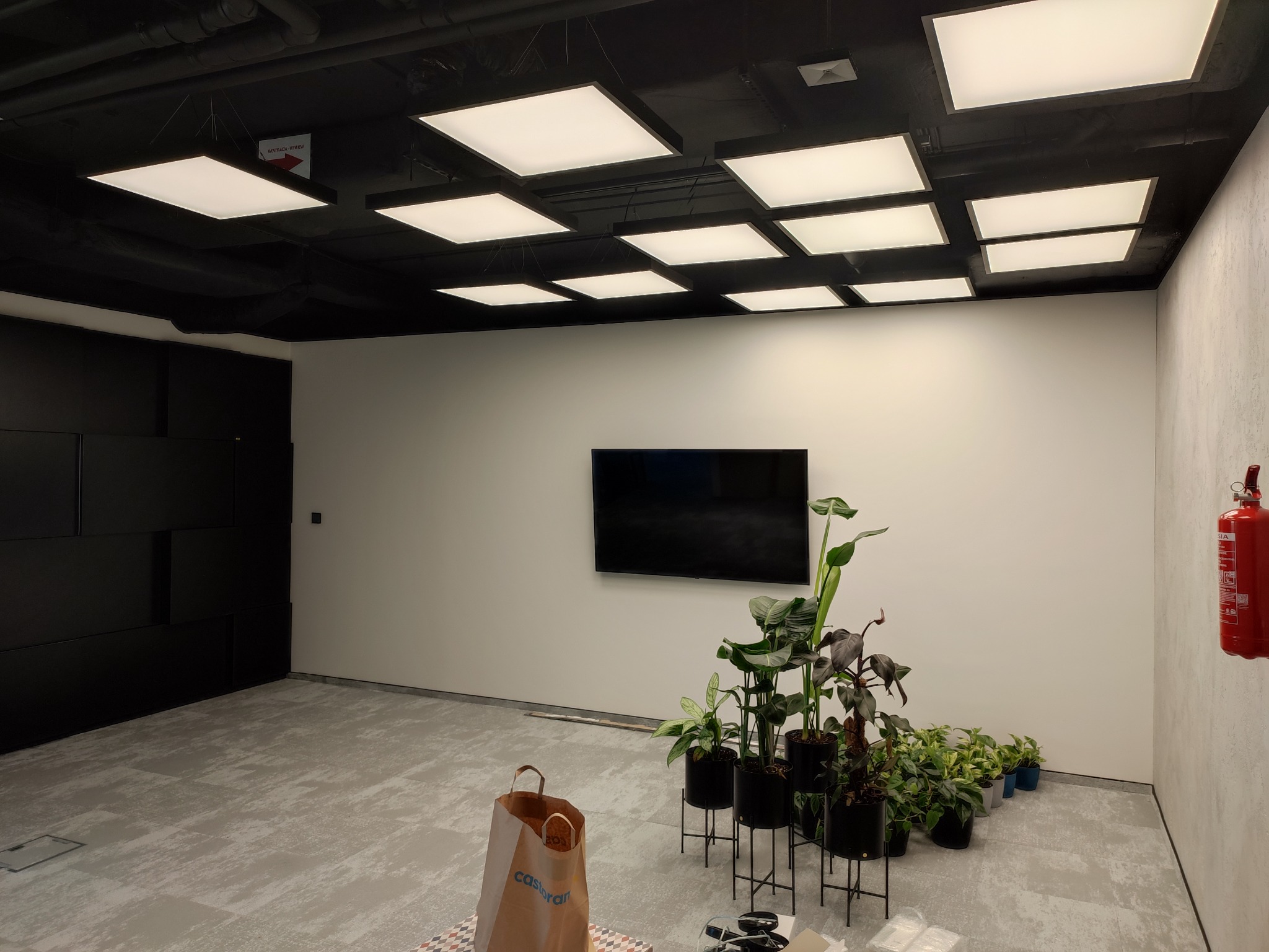 Biuro po montażu oświetlenia sufitowego LED, widoczne rośliny doniczkowe, telewizor na ścianie i gaśnica.