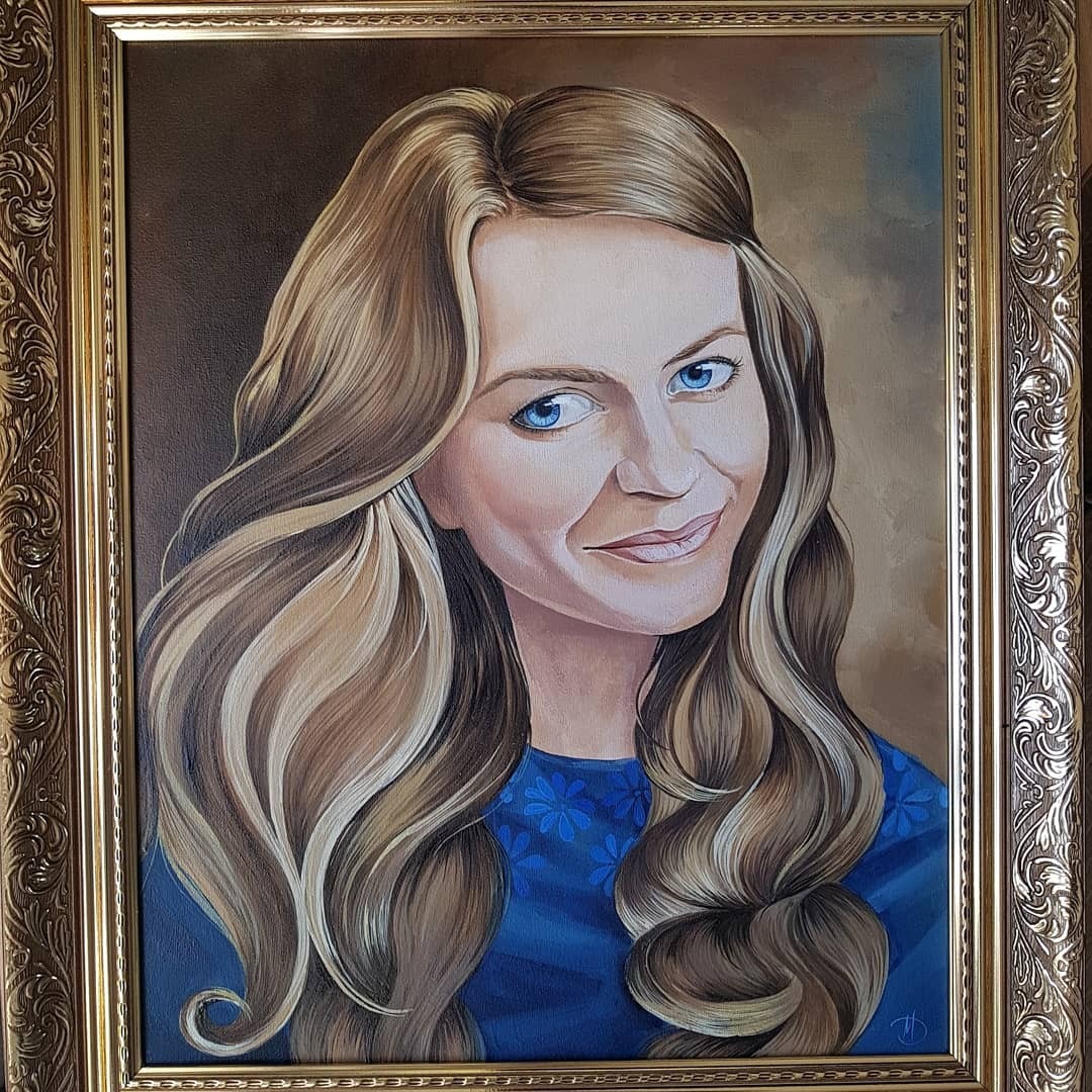 Portret olejny uśmiechniętej kobiety o blond włosach i niebieskich oczach, ubrana w niebieską bluzkę w kwiatki, oprawiony w bogato zdobioną, złotą ramę.