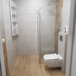 Nowoczesna łazienka z kabiną prysznicową typu walk-in, podwieszaną toaletą, grzejnikiem drabinkowym i drewnopodobną podłogą, wykończona płytkami w odcieniach szarości i drewna.