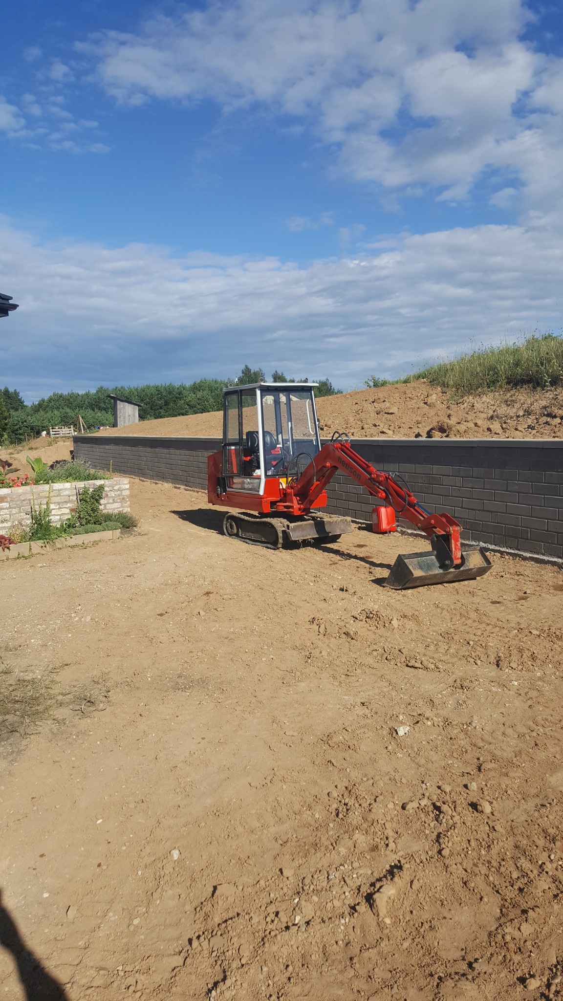 Czerwona mini koparka Neuson na piaszczystym terenie, widoczna szara ściana z bloczków betonowych i fragmenty zieleni w tle, błękitne niebo z chmurami.