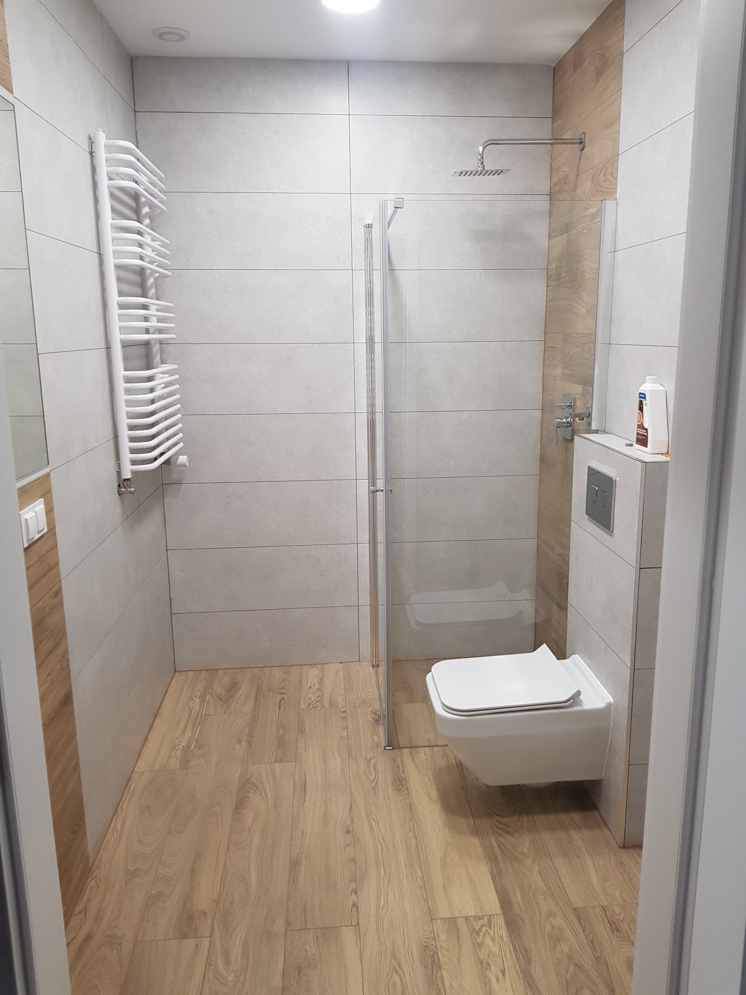 Nowoczesna łazienka z kabiną prysznicową typu walk-in, podwieszaną toaletą, grzejnikiem drabinkowym i drewnopodobną podłogą, wykończona płytkami w odcieniach szarości i drewna.