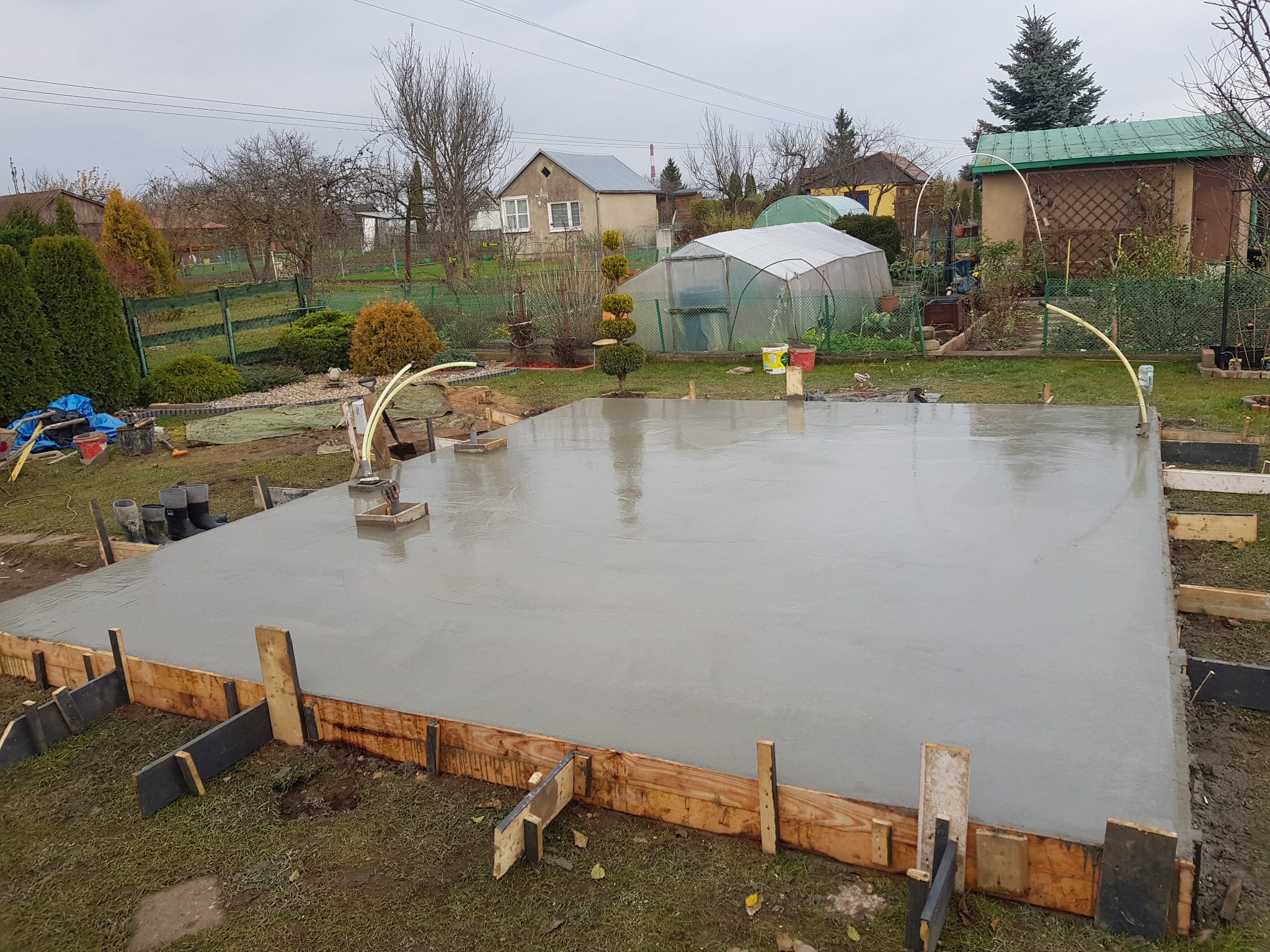 Świeżo wylana, mokra płyta fundamentowa otoczona drewnianym szalunkiem na trawiastym terenie działki z widocznymi budynkami mieszkalnymi i szklarnią w tle.