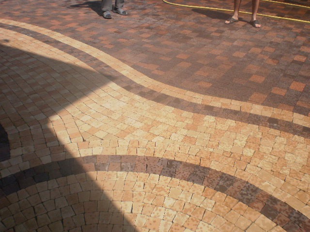 Ułożona kostka brukowa w odcieniach beżu i brązu, tworząca falisty wzór na podjeździe, częściowo zacieniona, z widocznymi stopami dwóch osób w tle.