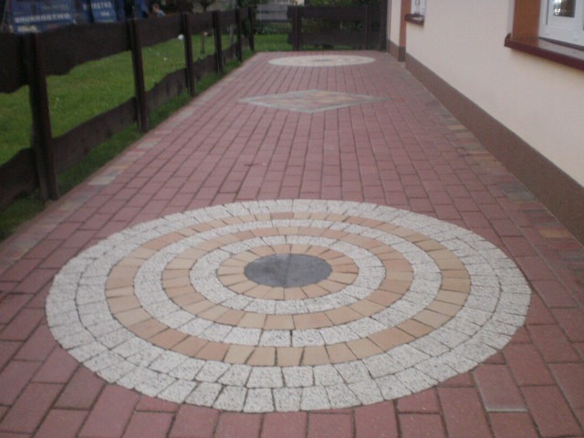 Ułożona kostka brukowa w kolorze ceglastym z dekoracyjnymi okręgami z jasnej kostki, widok z perspektywy chodnika wzdłuż budynku z drewnianym płotem.