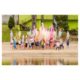 Kacper Polaczyk - Grupa dzieci i młodzieży na piaszczystej plaży rzuca w górę kolorowe proszki Holi, tworząc chmury barw w odcieniach różu, żółci, błękitu i zieleni, z budynkiem w tle.