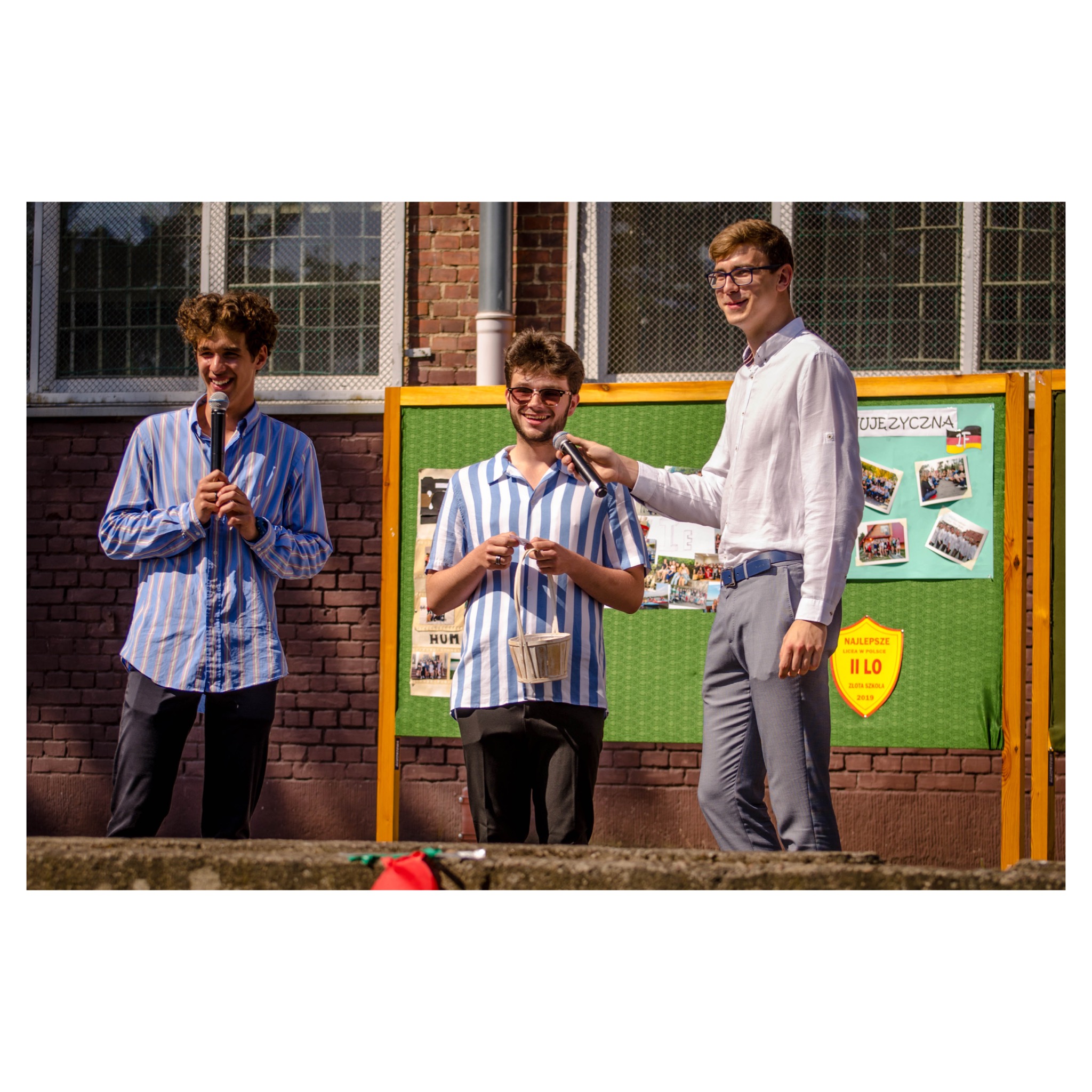Trzej młodzi mężczyźni w paski ubrani, stojący na scenie z mikrofonami, jeden trzyma wiklinowy koszyk, w tle tablica z napisem 'Ojczyzna' i zdjęciami.