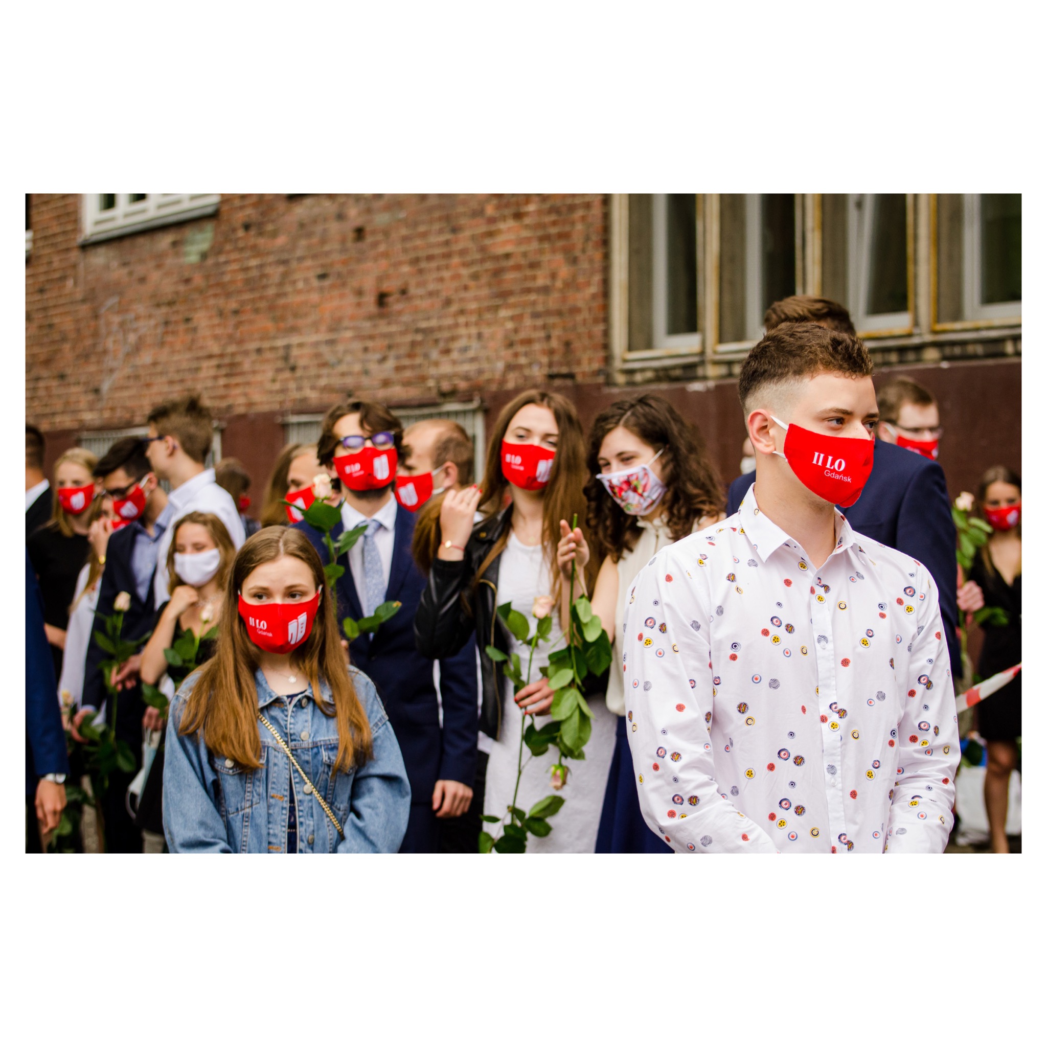 Grupa maturzystów w maseczkach z logo szkoły trzyma róże na tle ceglanego budynku.