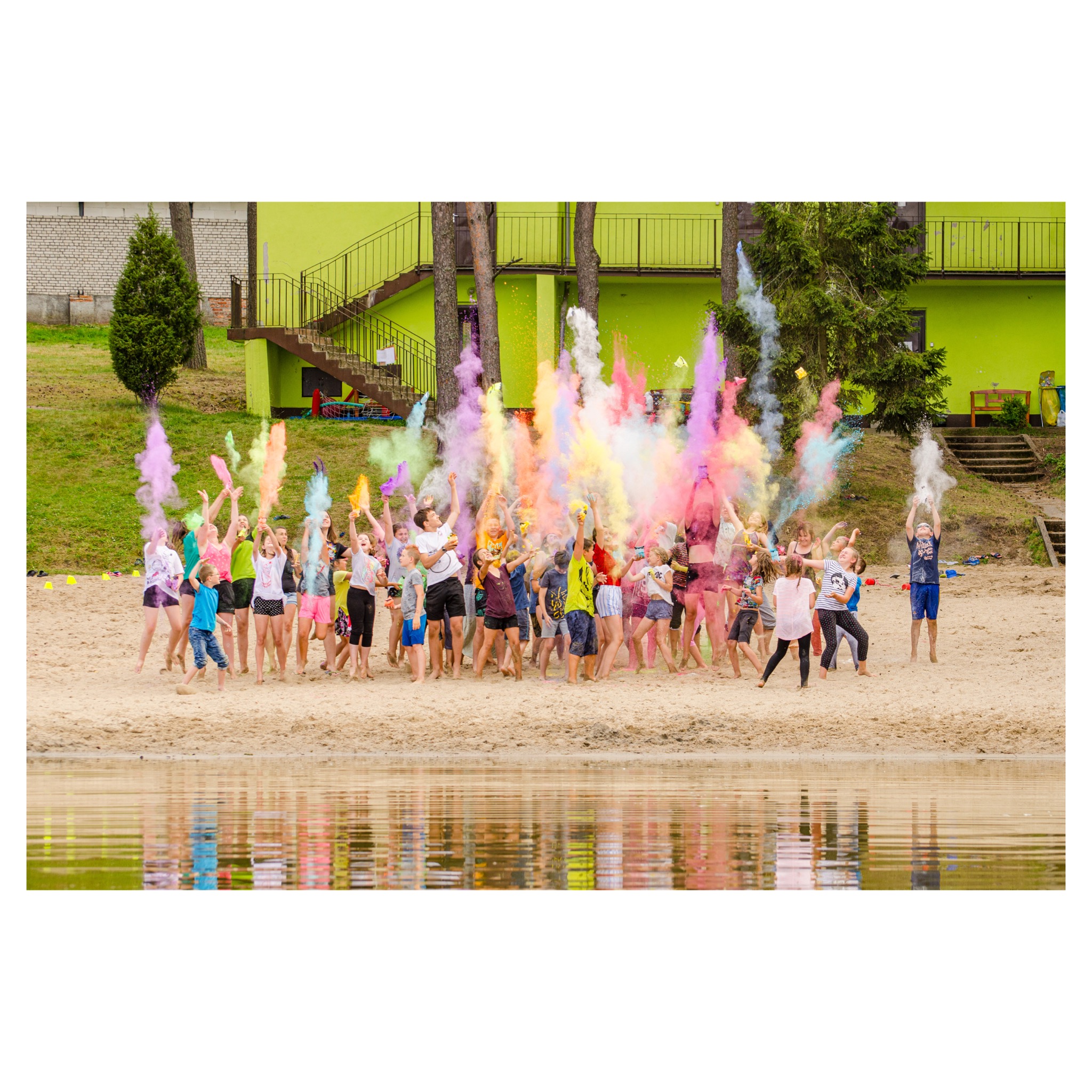 Grupa dzieci i młodzieży na piaszczystej plaży rzuca w górę kolorowe proszki Holi, tworząc chmury barw w odcieniach różu, żółci, błękitu i zieleni, z budynkiem w tle.