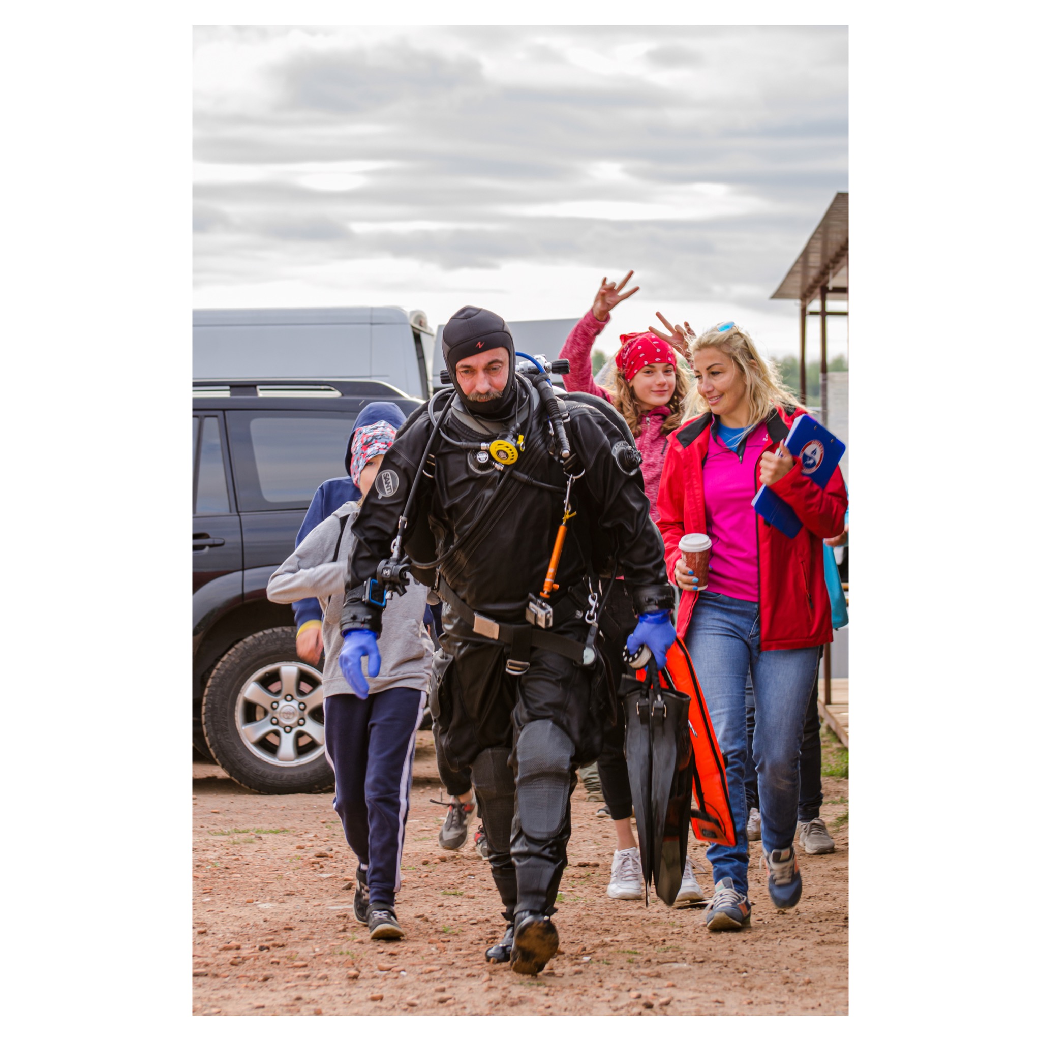 Nurek w pełnym kombinezonie, z płetwami i sprzętem, idzie w grupie osób po piaszczystym terenie, w tle zaparkowany samochód.