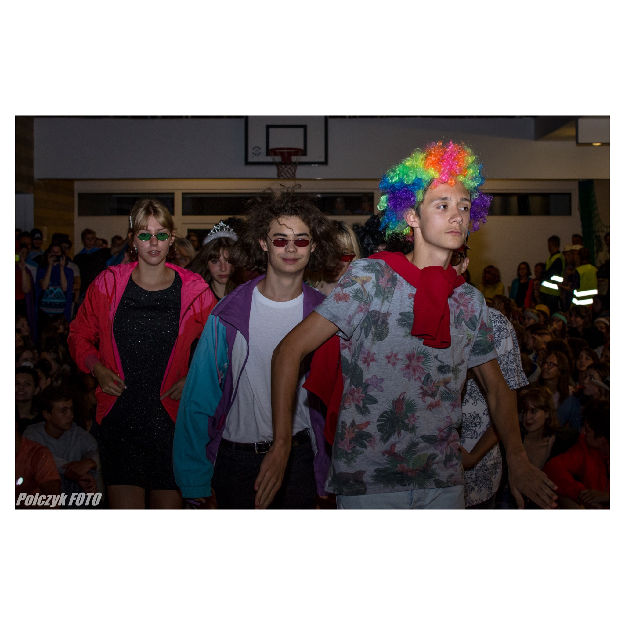 Młodzież w barwnych strojach i perukach, prawdopodobnie podczas szkolnego przedstawienia lub imprezy tematycznej, na tle widowni i boiska do koszykówki.