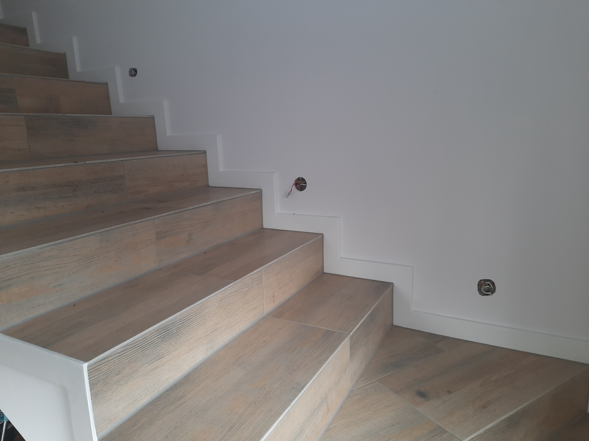 Schody wyłożone płytkami imitującymi drewno, z białymi listwami przypodłogowymi i przygotowanymi otworami na oświetlenie w ścianie.