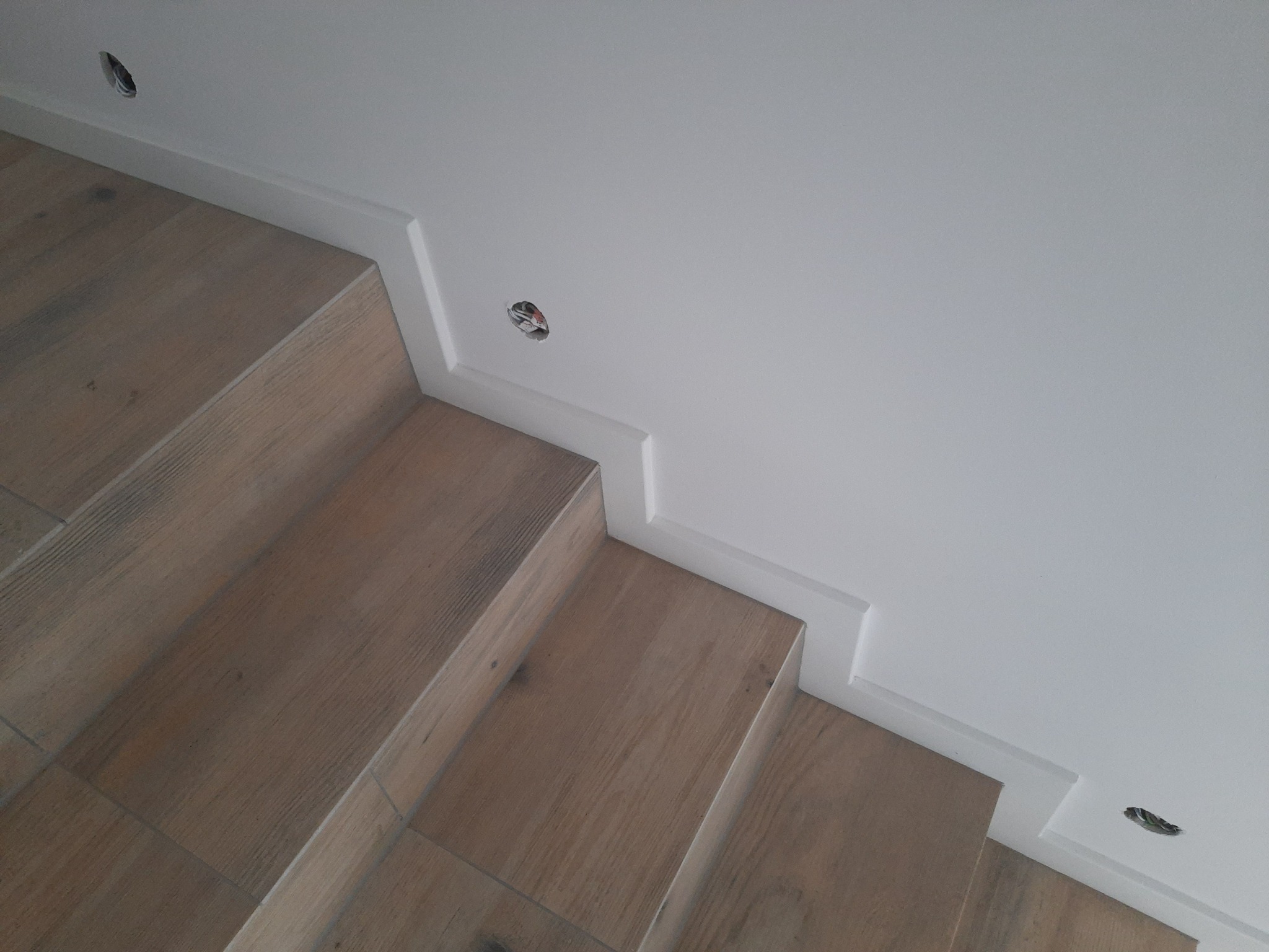 Ukończone schody z drewnopodobnych płytek, biała ściana z ozdobną listwą przysufitową i otworami na oświetlenie punktowe.