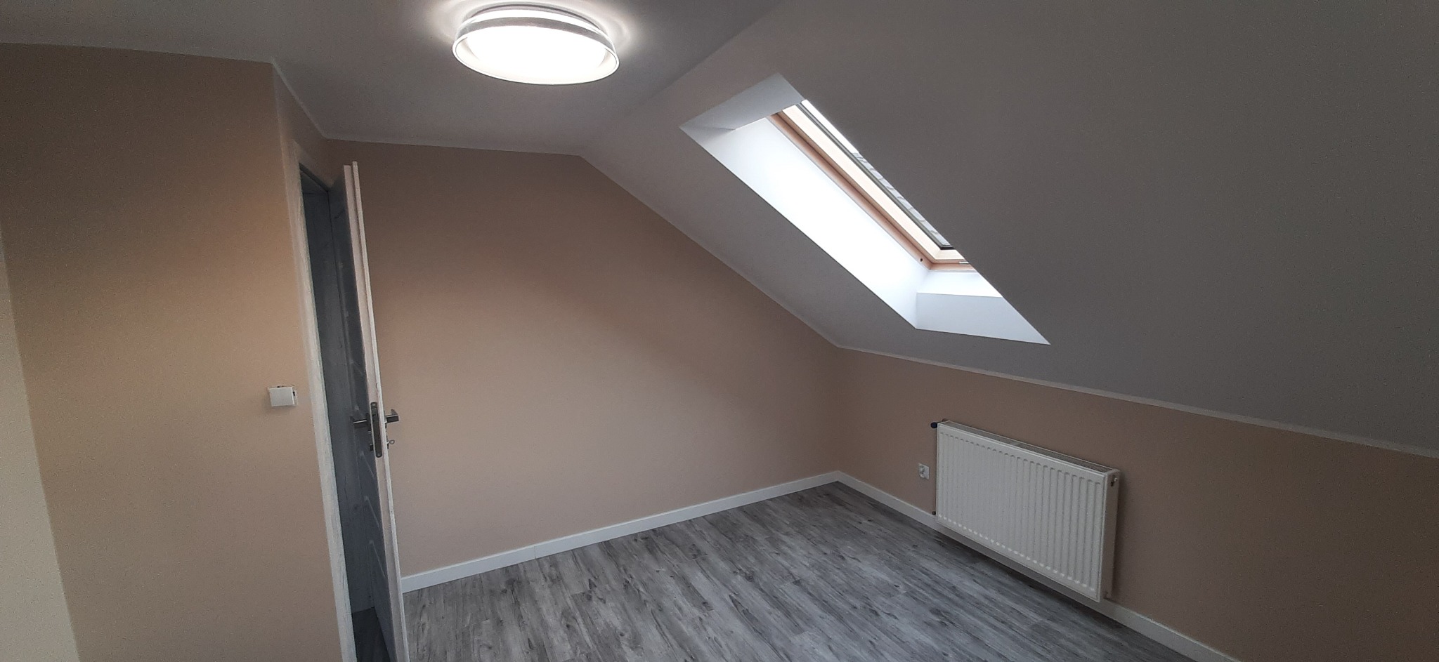 Poddasze po remoncie: beżowe ściany, szara podłoga imitująca drewno, biały grzejnik, okno dachowe i okrągła lampa sufitowa.