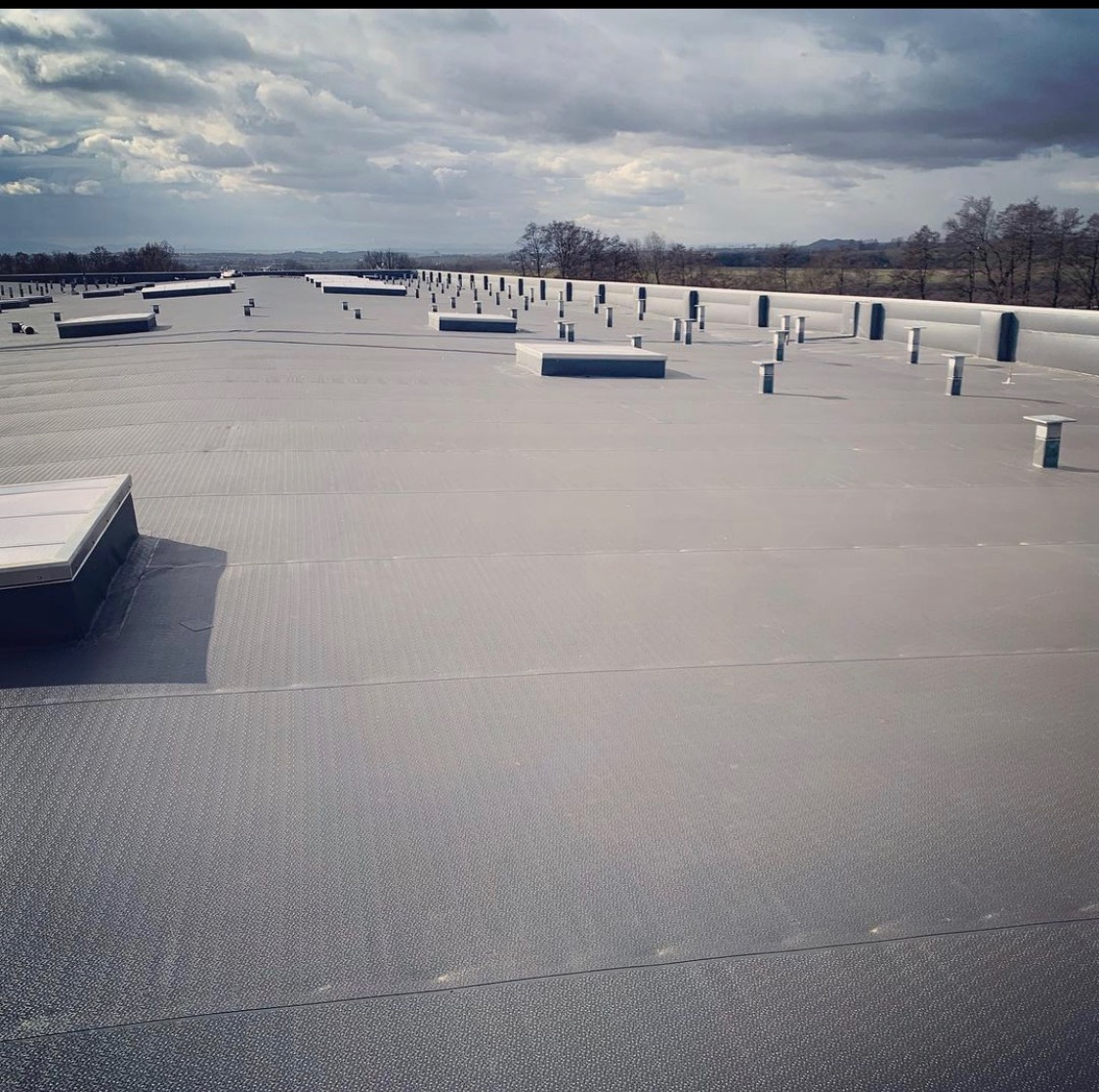 Rozległy, płaski dach pokryty ciemną membraną, z licznymi kominkami wentylacyjnymi i świetlikami, rozciągający się w kierunku odległego krajobrazu pod pochmurnym niebem.