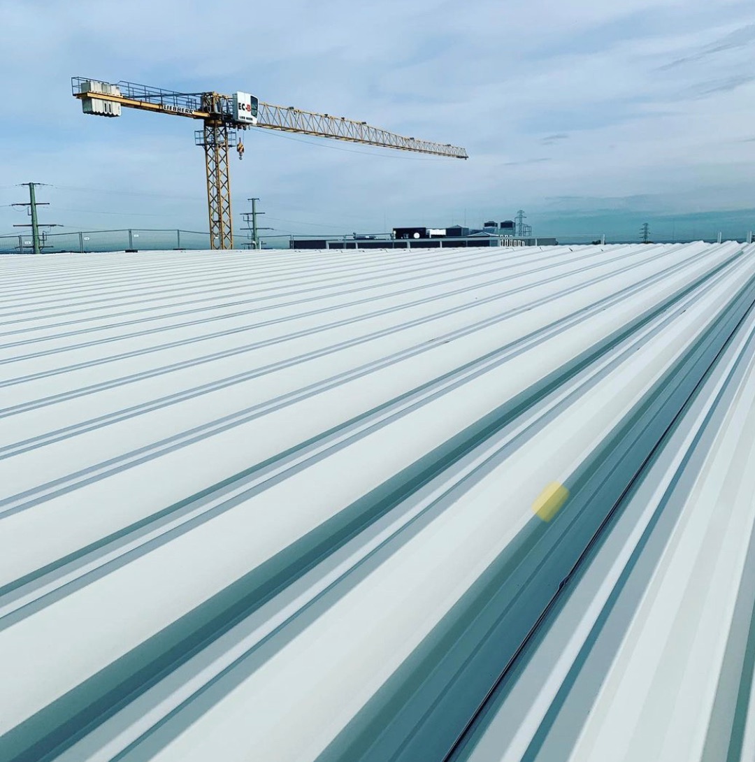 Widok z góry na nowo ułożone, białe arkusze blachy na dachu przemysłowym, z żółtym dźwigiem budowlanym i liniami wysokiego napięcia w tle pod błękitnym niebem.