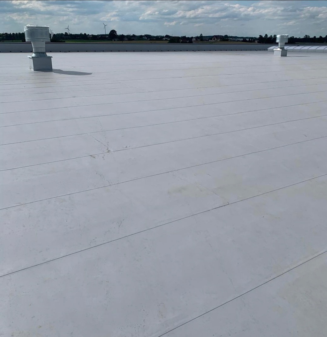 Jasnoszara, płaska powierzchnia dachu przemysłowego z widocznymi łączeniami, wentylatory dachowe i odległy krajobraz z turbinami w tle.