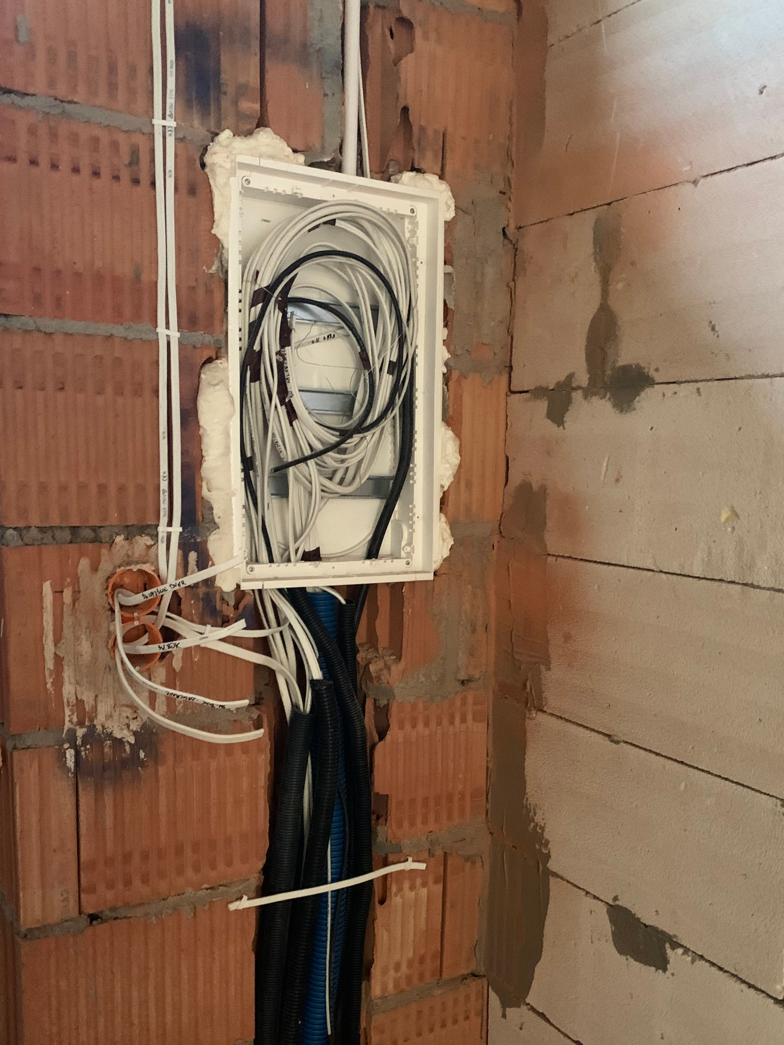 Rozdzielnica elektryczna w stanie surowym, z widocznymi przewodami w peszlach i bez, zamontowana w ścianie z czerwonej cegły, uszczelniona pianką montażową.