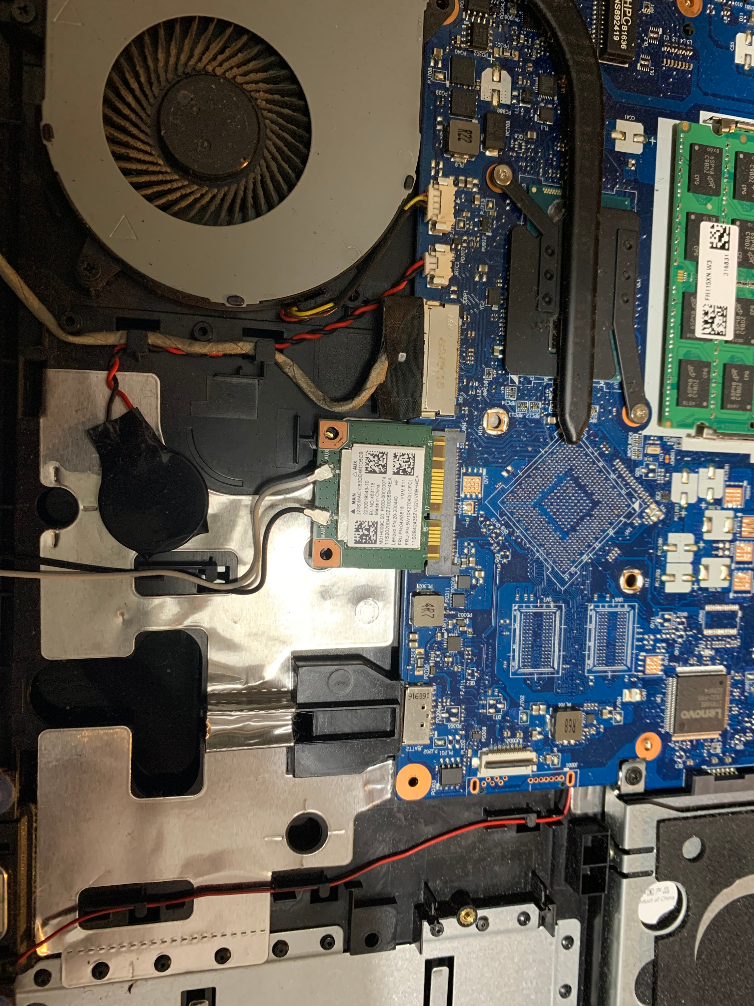 Wnętrze laptopa przed czyszczeniem: widoczny zakurzony wentylator chłodzenia, płyta główna z elementami elektronicznymi, bateryjka podtrzymująca BIOS oklejona czarną taśmą oraz karta wifi.