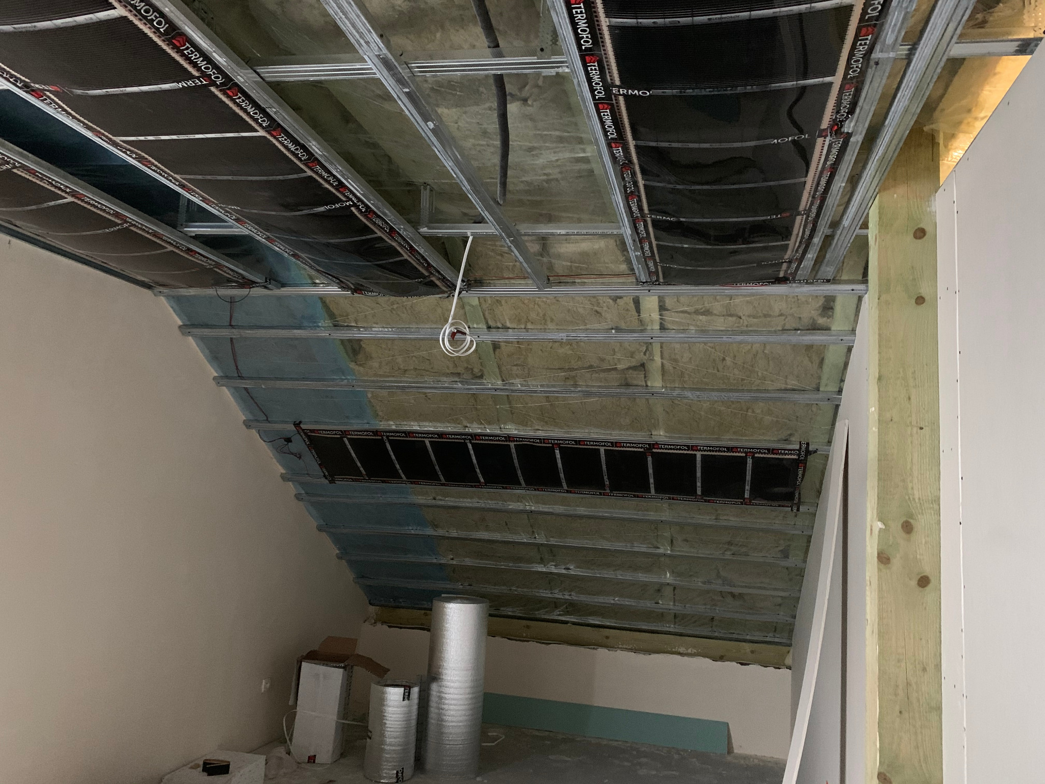 Montaż mat grzewczych Termofol na poddaszu, widoczne profile stalowe, izolacja wełną mineralną i rolki materiału izolacyjnego w pomieszczeniu ze skosami.