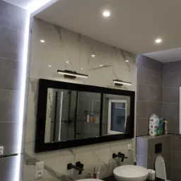 Nowoczesna łazienka z dwoma białymi umywalkami nablatowymi, czarną armaturą i dużym lustrem w czarnej ramie z oświetleniem LED. Na ścianach płytki imitujące marmur i szare płytki. Widoczne...